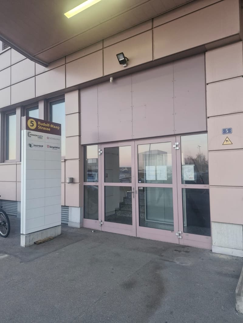 Building with entrance door, glass doors, sign with text '5 Rudolf-Maag-Strasse', security camera, bicycle