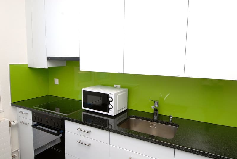 modern kitchen with green splashback, white cabinetry, black countertops, electric stove, oven, stainless steel sink, faucet, white microwave, power outlet