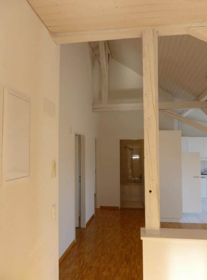 Empty room with wooden floor, wooden ceiling beam, white walls, doorways, and a shower area.