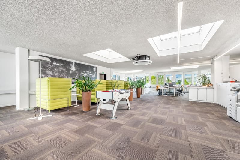 Bright and open office space with large windows, skylights, and a modern, minimalist design. The space features a yellow sofa, potted plants, and a foosball table, creating a relaxed and collaborative atmosphere. The flooring is a patterned carpet tile, a