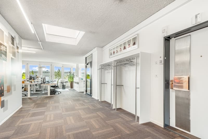 Spacious open-plan office with large windows, modern furniture, and a kitchenette area with a dishwasher