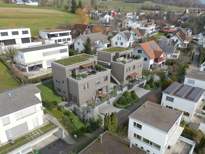 Multi-story apartment building with balconies, raised ground floor, and parking spaces. The building has a modern, minimalist design with a mix of white and gray colors. The surrounding area appears to be a residential neighborhood with other houses and b
