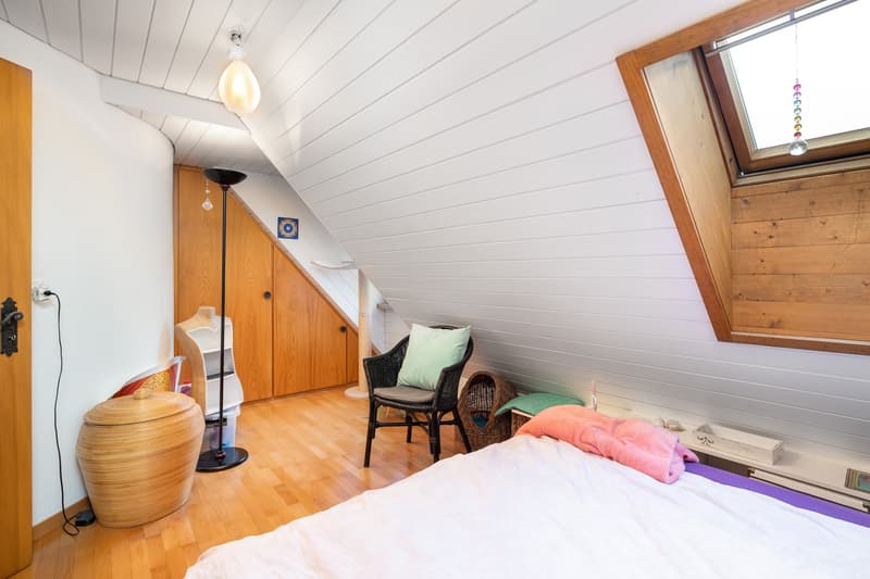 a cozy attic bedroom with white ceiling, wooden floor, a single bed, a chair, a bedside table, a floor lamp, a wooden storage bin, a wardrobe, and a skylight window