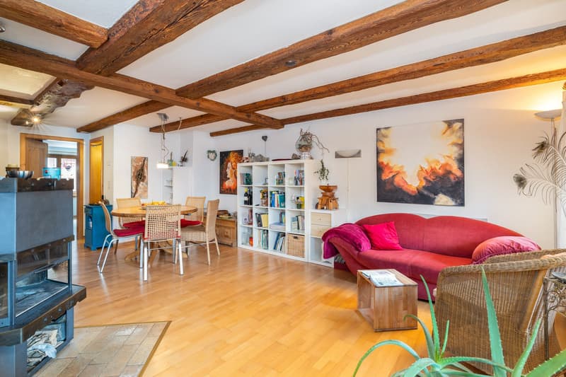 Fireplace, wooden flooring, wooden beams, red couch, dining table with chairs, bookshelf, paintings, wooden coffee table