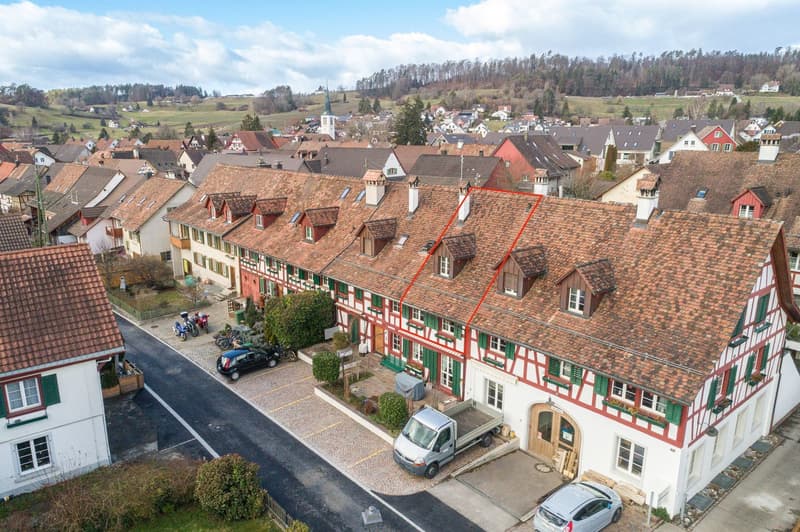 row house, brown roof, white walls with red accents, green shutters, multiple cars parked in front, small garden, chimneys