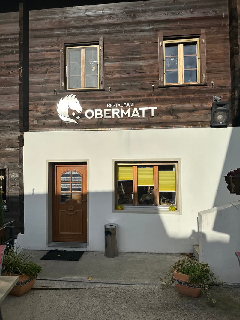 The image shows the exterior of a restaurant called 'Restaurant Obermatt'. The building has a wooden facade with large windows and a wooden door. The restaurant's logo is prominently displayed on the front of the building.