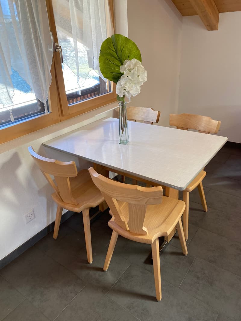 The image shows a dining room with a square wooden table and four wooden chairs. The table has a white marble-like top. There is a large window with curtains, providing natural light. A vase with a large green leaf and white flowers is placed on the table