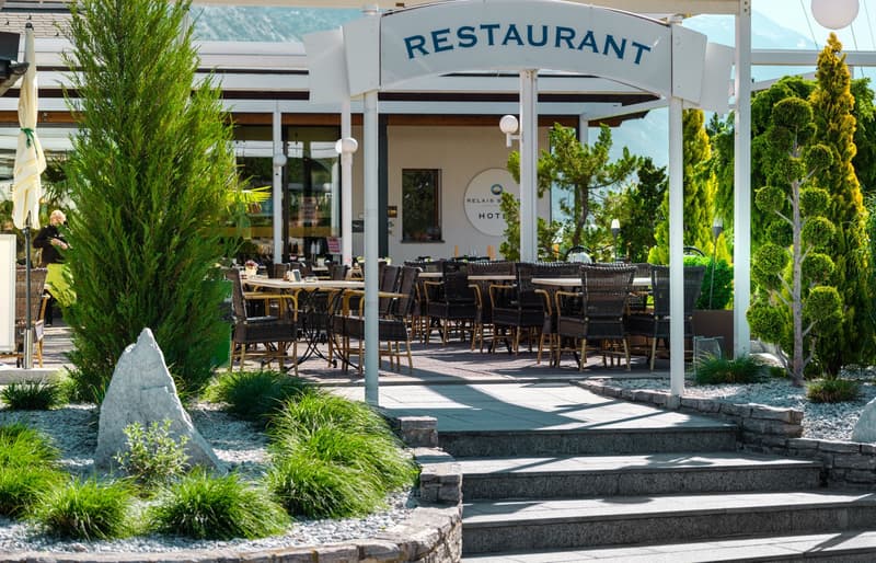 Outdoor restaurant seating area with tables and chairs, surrounded by lush greenery and landscaping. The restaurant is located at the Relais de Margaux Hotel, as indicated by the sign.