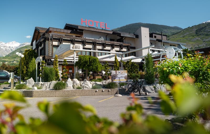 A large hotel building with a prominent 'HOTEL' sign on the roof, surrounded by lush greenery and mountains in the background. The hotel has a terrace and outdoor seating area, and there is a bicycle rack in the foreground.