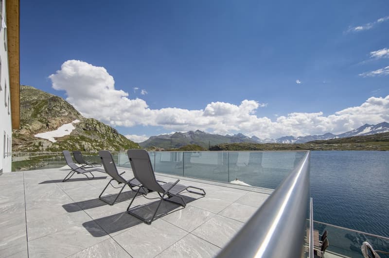 Spacious outdoor terrace with glass railing, overlooking a scenic lake surrounded by mountains. Three lounge chairs are set up on the terrace, providing a comfortable spot to enjoy the stunning natural views.