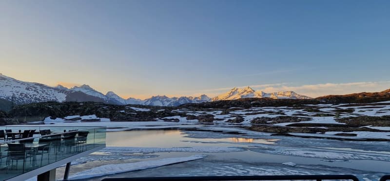The image shows a stunning winter landscape with snow-capped mountains in the background. In the foreground, there is a frozen lake or pond with a glass-enclosed structure, possibly a restaurant or hotel, overlooking the scenic view. The sky is a beautifu