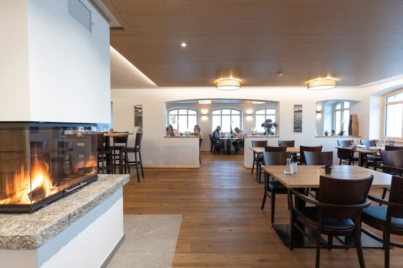 Spacious dining area with wooden floors, arched windows, and a fireplace. Tables and chairs are arranged throughout the room, and there are people seated at some of the tables. The lighting is warm and inviting, with drum-shaped light fixtures hanging fro