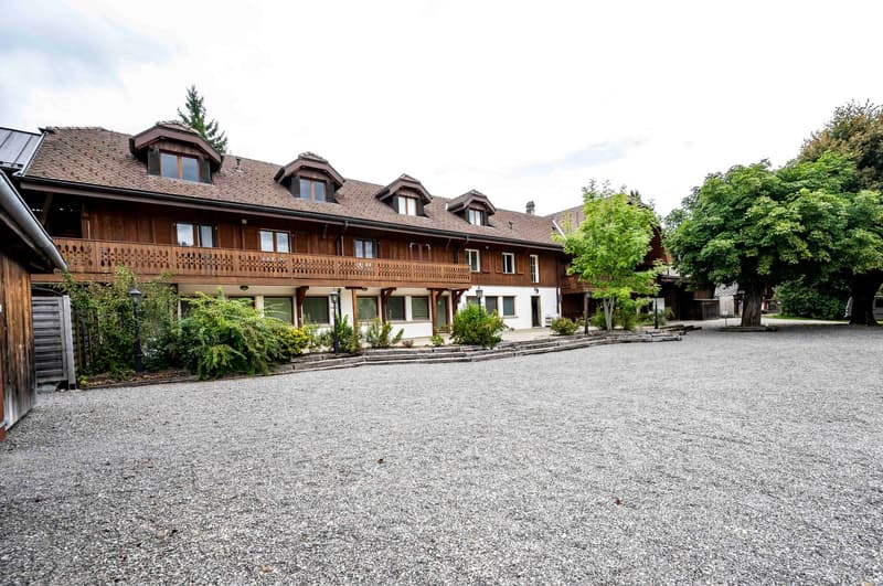 Wooden building with balconies, gravel driveway, several windows, surrounded by trees and shrubs