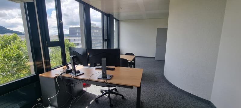 Spacious office with large windows providing a view of the surrounding buildings and greenery, desk with multiple computer monitors, office chair, and additional seating area