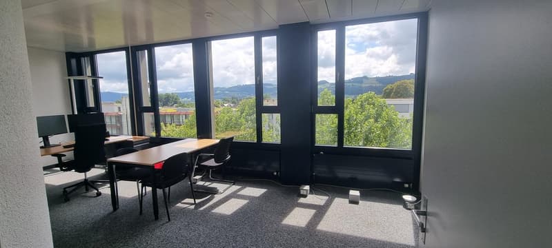 Large office space with floor-to-ceiling windows providing a scenic view of the surrounding greenery and hills. The room has a modern, minimalist design with black furniture and equipment, including desks, chairs, and computer monitors.