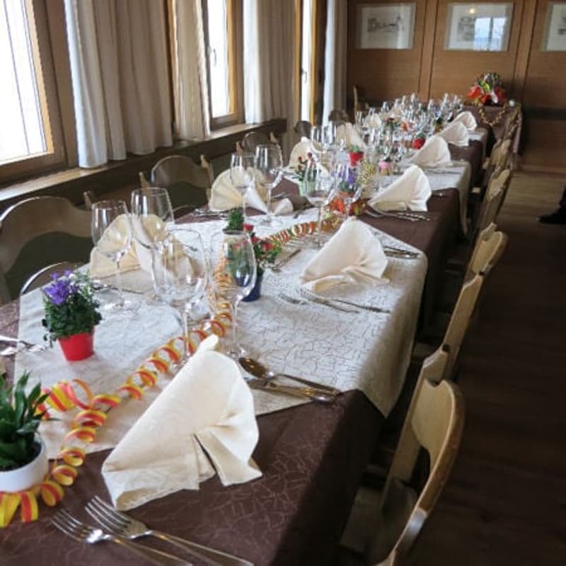 long dining table, set for multiple people, wine glasses, silverware, napkins, flowers, wooden chairs, wooden walls, windows with curtains