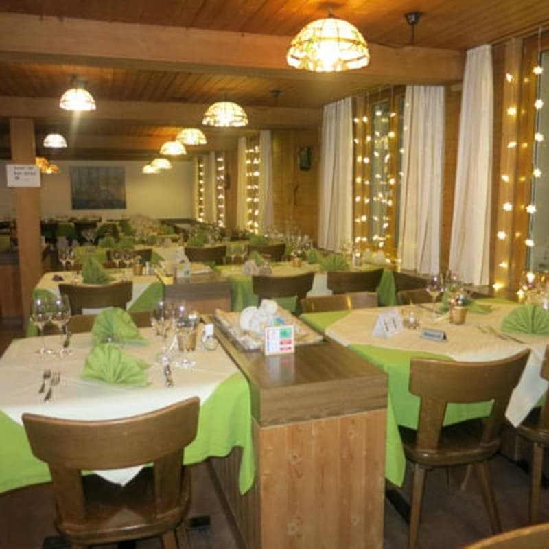 Restaurant with wooden ceiling, white curtains, wooden tables and chairs, green table cloths, wine glasses, forks, knives, and spoons