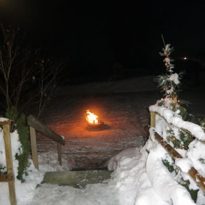 Outdoor fireplace in the snow, surrounded by wooden fences, steps leading to the fireplace, Christmas tree with lights