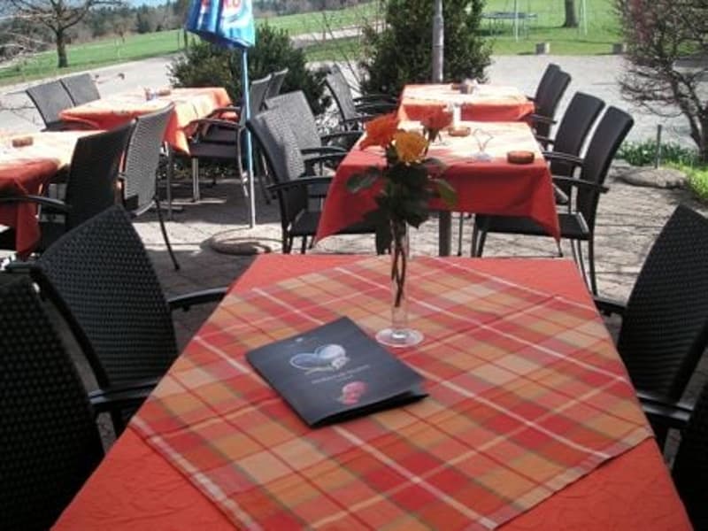 Outdoor dining area with tables and chairs, red tablecloths, flowers in vases, umbrella, menu on the table, view of a golf course