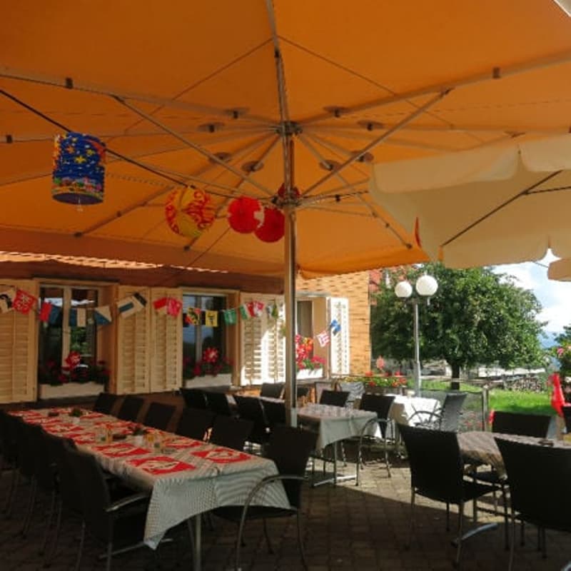 Outdoor dining area with tables and chairs, large umbrellas, flags, and a building in the background