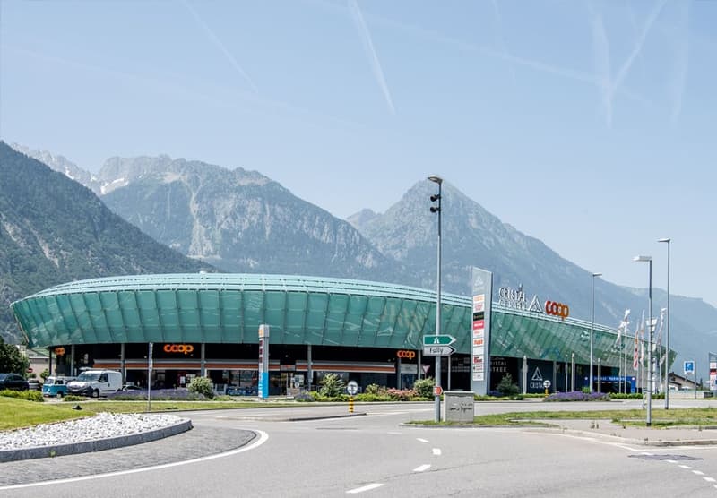Large oval building with a green facade, several cars parked in front, sign with white arrow, mountains in the background