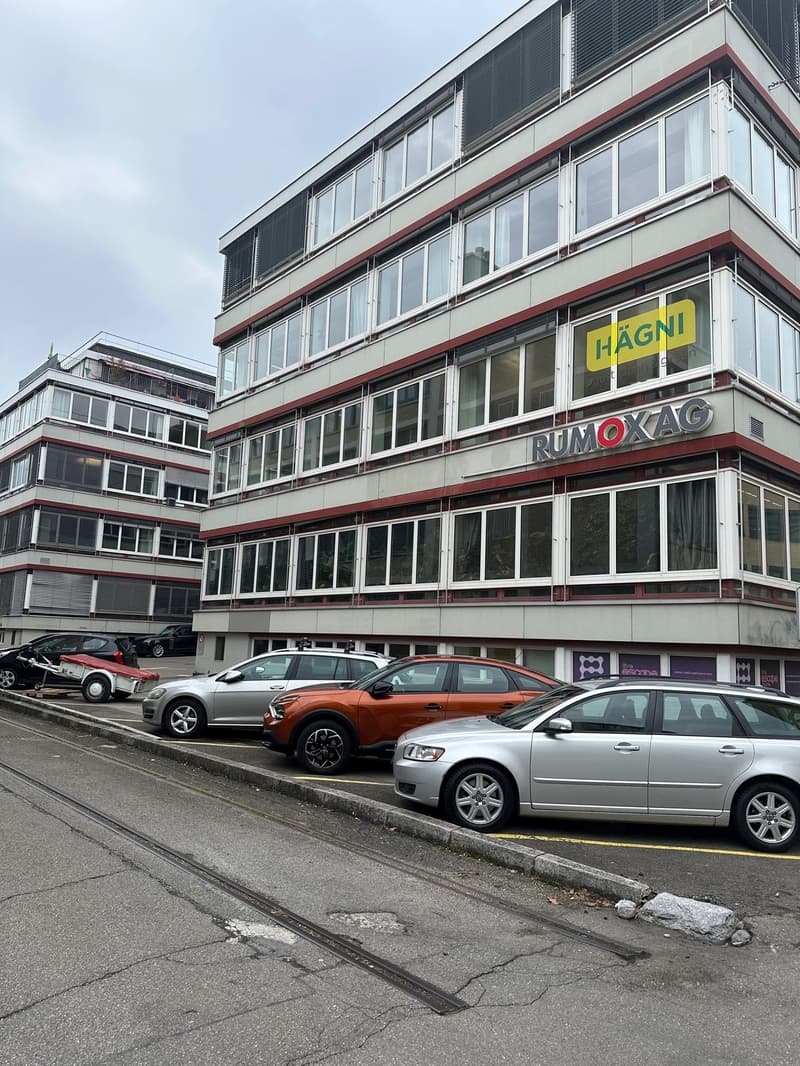 A multi-story building with several parking spaces on the front. The building has red brick walls and white windows. The parking space has several cars parked on it. The street in front of the building has a tram track on the left. There are two other bui