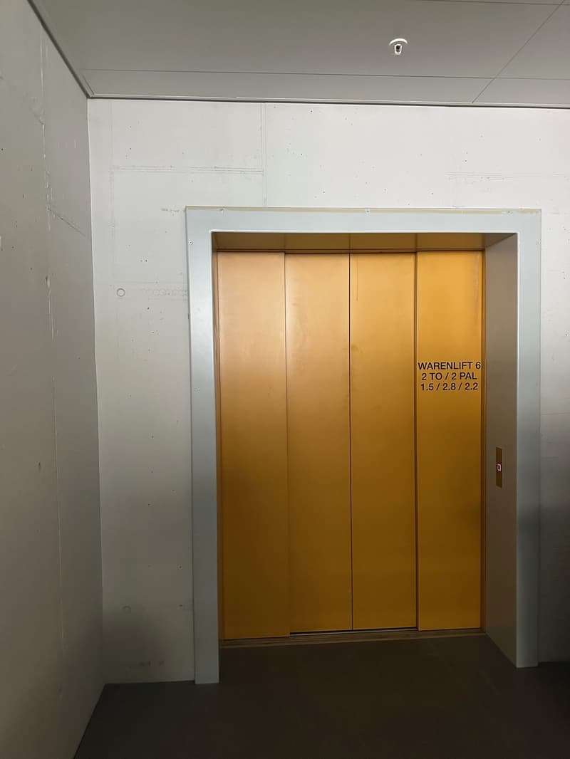 Empty elevator shaft with white walls and yellow doors, elevator labeled 'WARENLIFT 6 2 TO/2 PAL 1.5/2.8/2.2'