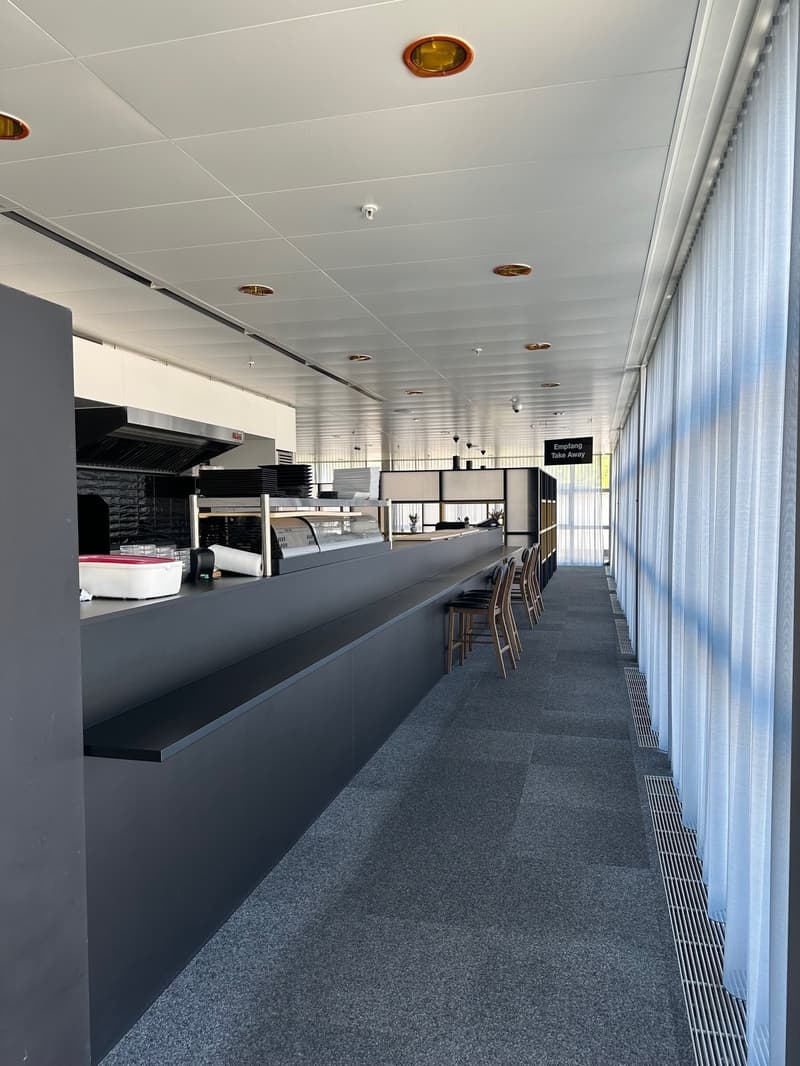 A counter with stacks of plates and food containers, black walls, gray flooring, a row of chairs, and ceiling lights.