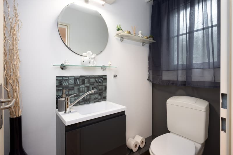 modern bathroom with white walls, black cabinet, white sink, silver faucet, round mirror, glass shelf, toilet, toilet paper, window with curtain
