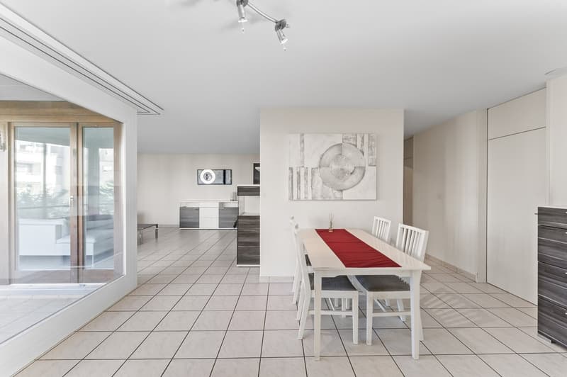Minimalist dining room with white tiles, a large table, a white chair with a gray seat, and a glass door with a balcony view outside.