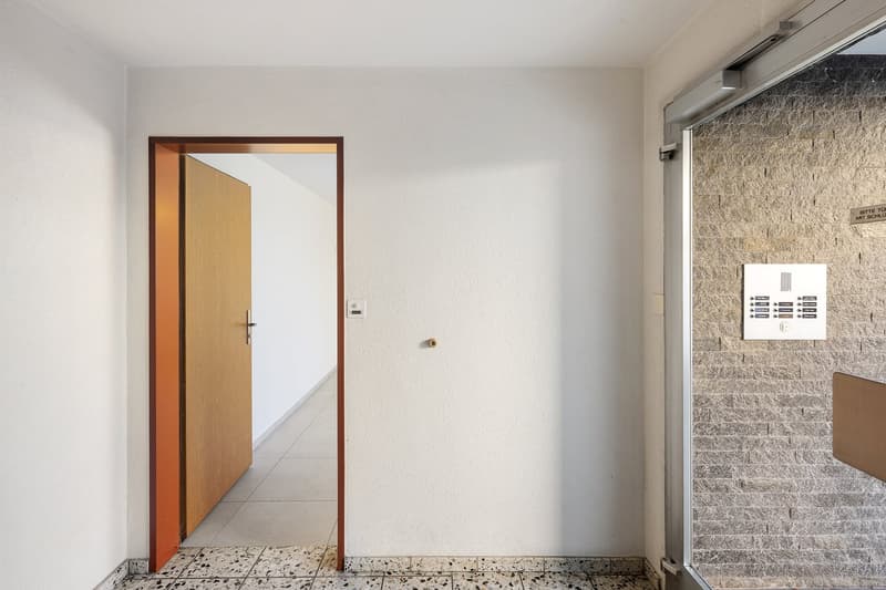 Empty hallway with white walls, wooden door, tiled floor, and an open door leading to another room.