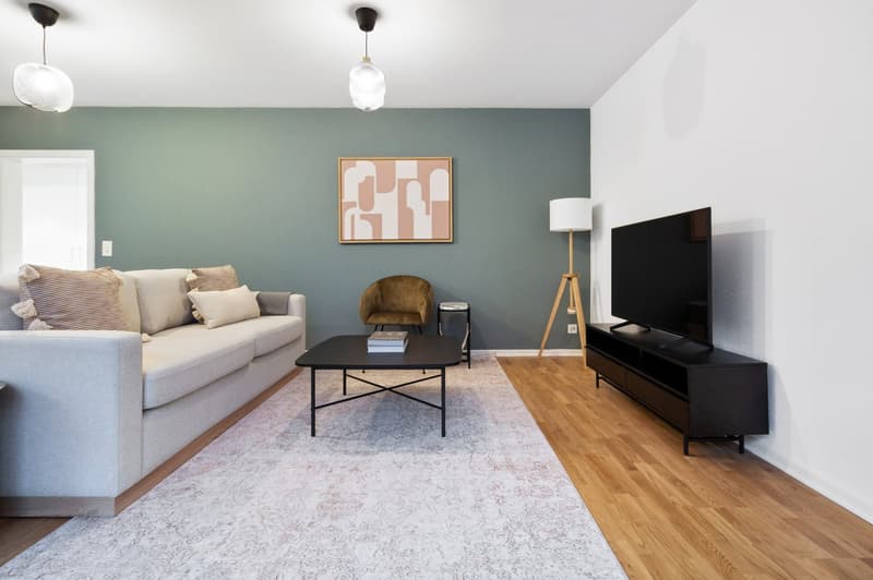Spacious living room with gray sofa, wooden coffee table, black TV stand, hardwood floors, and abstract artwork on the wall. The room also features a floor lamp and a pendant light fixture.