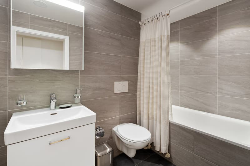Modern bathroom with gray tile walls, white vanity with sink, toilet, bathtub, and shower curtain