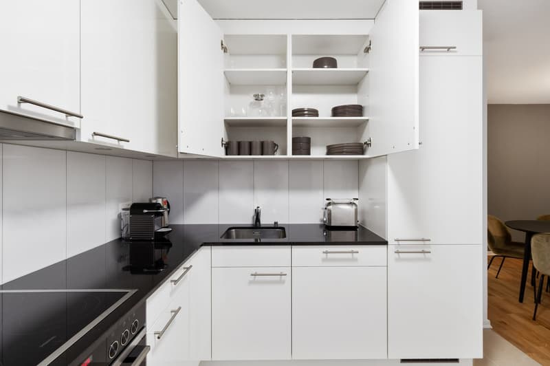 Modern, minimalist kitchen with white cabinets, black countertops, built-in appliances including a dishwasher and coffee machine, and open shelving displaying dishes and cookware.