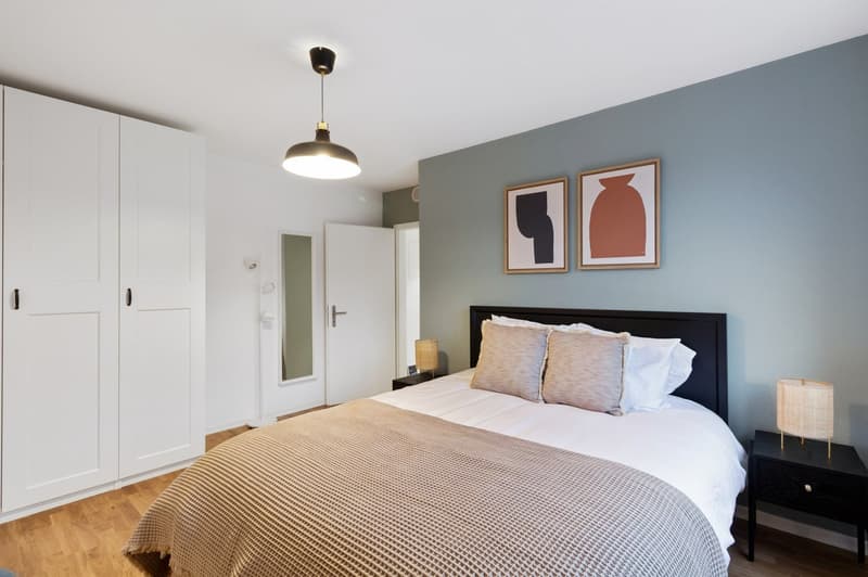 Spacious bedroom with white built-in wardrobes, gray walls, large bed with beige bedding, abstract artwork, and a pendant light fixture.