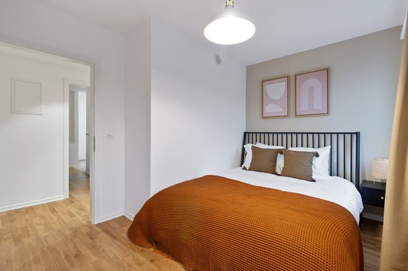 Spacious bedroom with white walls, hardwood flooring, a large bed with an orange blanket, and two framed artwork pieces on the wall. The room has a simple and modern design.