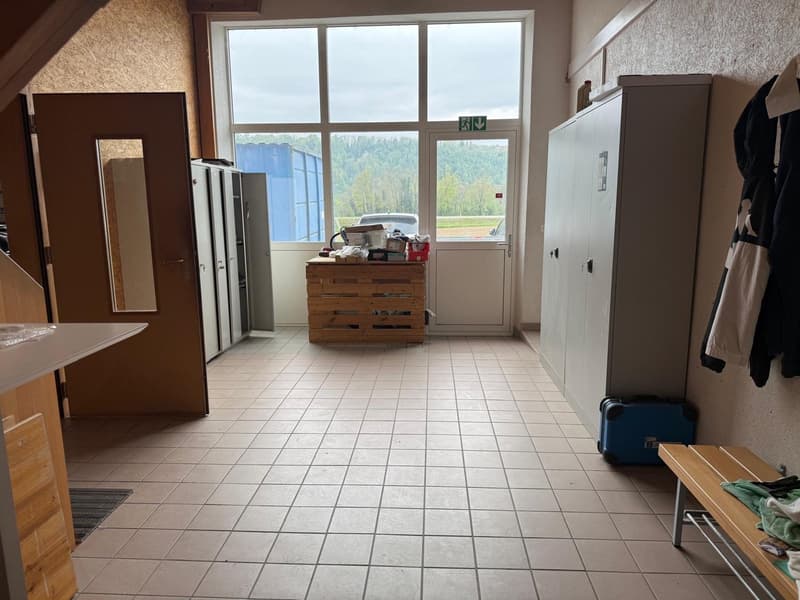 This image shows a room with a large window overlooking a scenic outdoor view. The room has tile flooring and contains various furniture and appliances, including a refrigerator, cabinets, and a wooden crate used for storage.