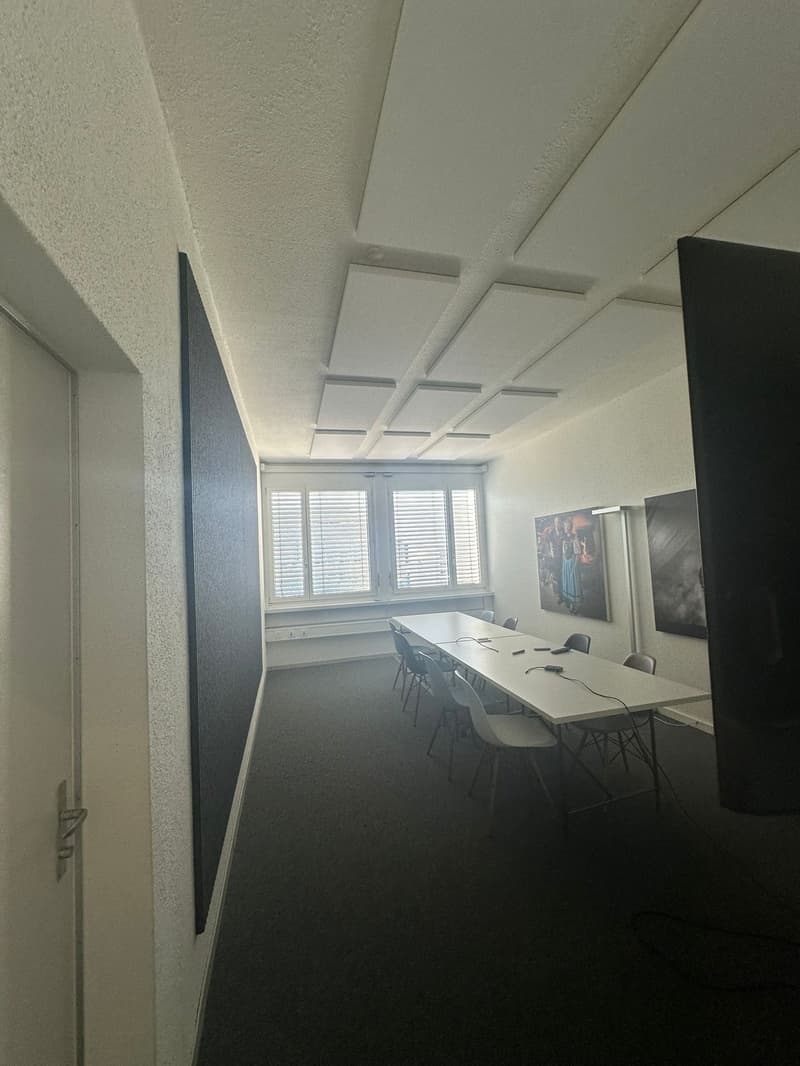 The image shows a modern office space with a large conference table, chairs, and windows with blinds. The ceiling has a grid-like pattern with recessed lighting. The walls are painted a light color, and there is artwork displayed on one of the walls.