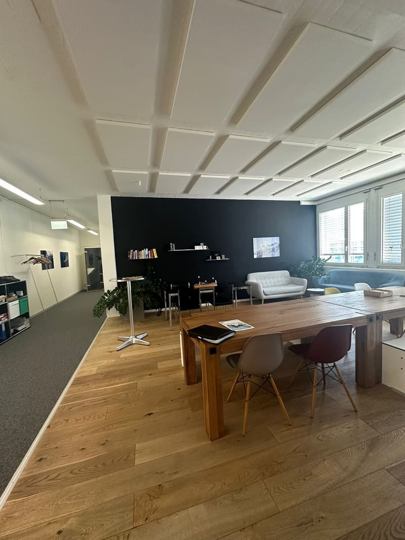 Open concept office space with hardwood floors, white walls, and a coffered ceiling. The space includes a large wooden table with chairs, a sofa, and shelving units on the back wall. There is also a dishwasher visible in the room.
