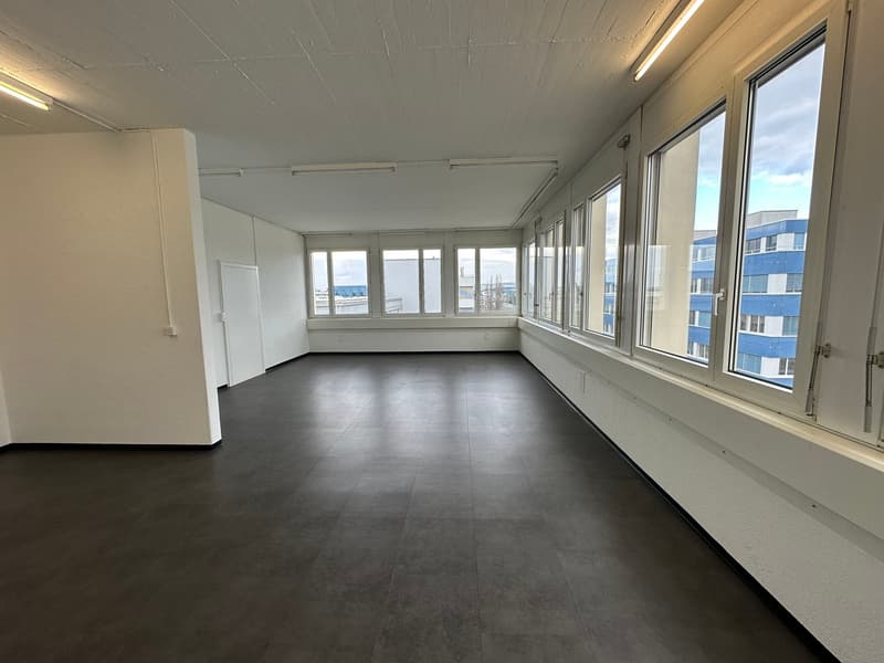 Wide and open room with windows and view to the outside, dark floor, white walls, 4 light sources on the ceiling.
