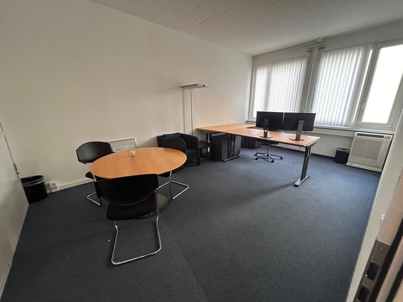 A large room with blue carpet flooring, two round tables, one of which has a candle, two computers on desk, four chairs, one lamp in the corner, three windows, two trash bins, a power outlet on the wall