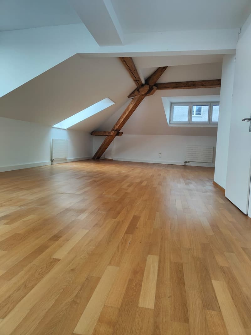 Empty attic with wooden beams, hardwood floors, white walls, two attic windows, a ceiling radiator on each side of the room.