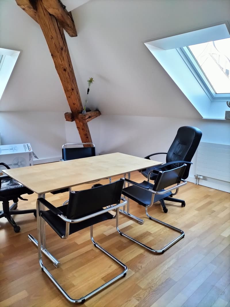 Attic office space with wooden beams, large windows, wooden table, black office chairs, and a small plant on the table.