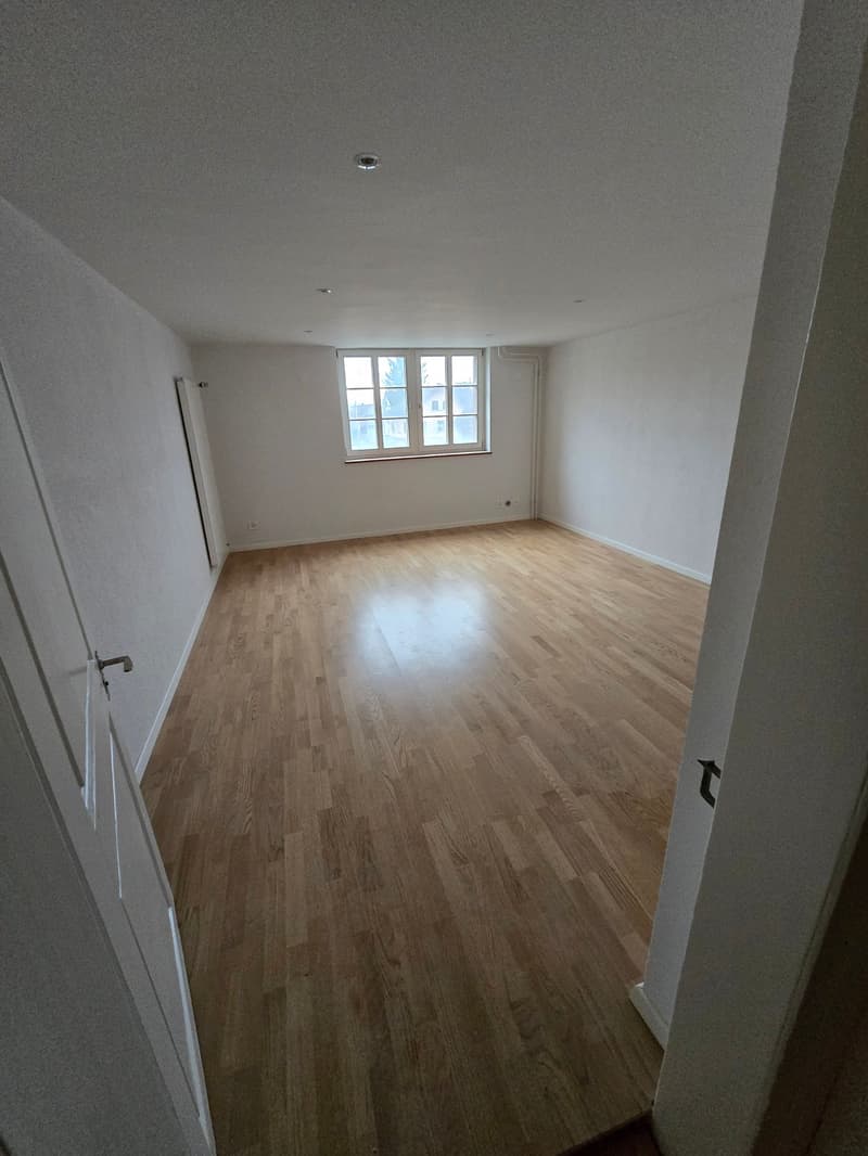 Empty room with hardwood floor, white walls, large window, door.