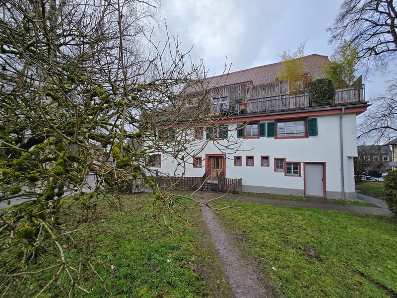 White building with red accents, wooden roof, balcony, garage, surrounded by a lawn and trees.