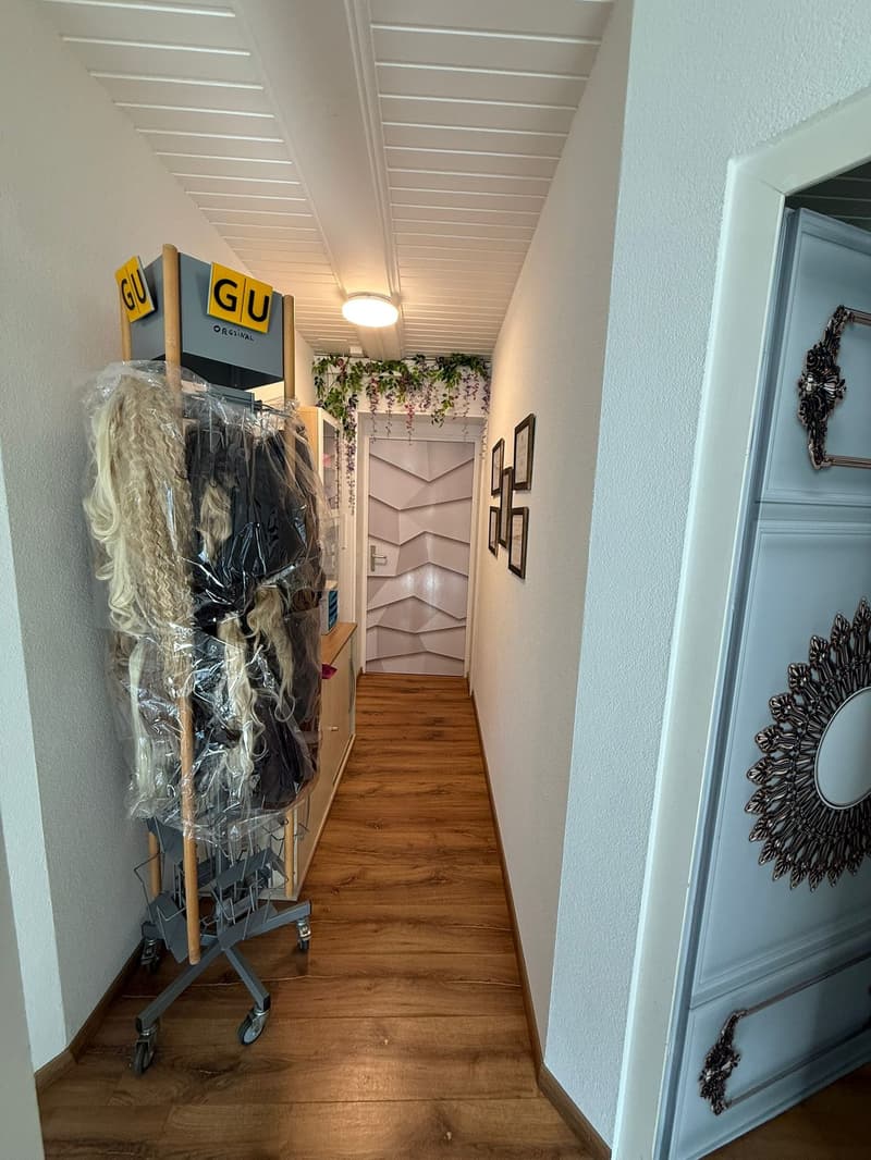 Corridor with wooden floor, white ceiling, racks with wigs, storage boxes, wall-mounted mirrors, framed pictures, light fixture, and an entrance to a room.