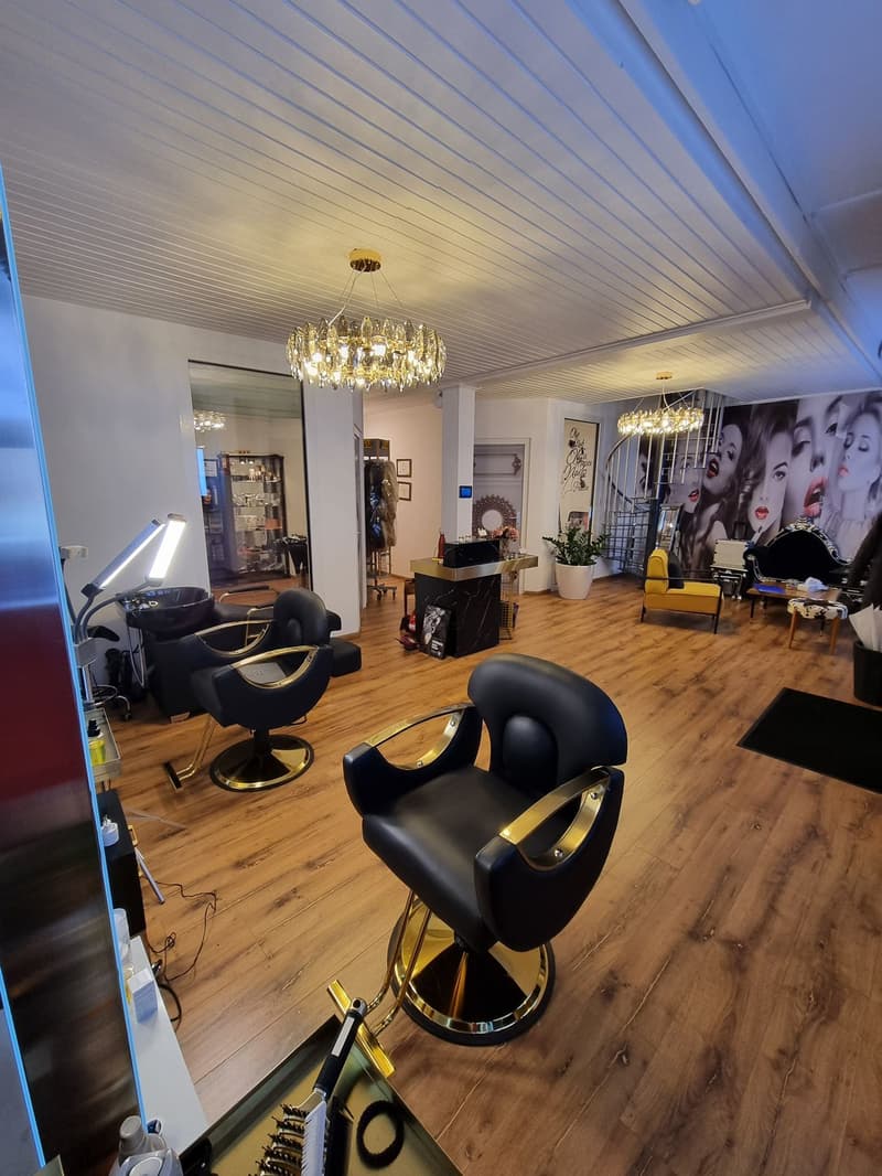 A salon with a wooden floor, a chandelier, two black swivel chairs, and a desk with a potted plant. The walls have large mirrors, and the room has a white ceiling with a pattern of lights. A large picture of women is on the wall. There is also a lamp and