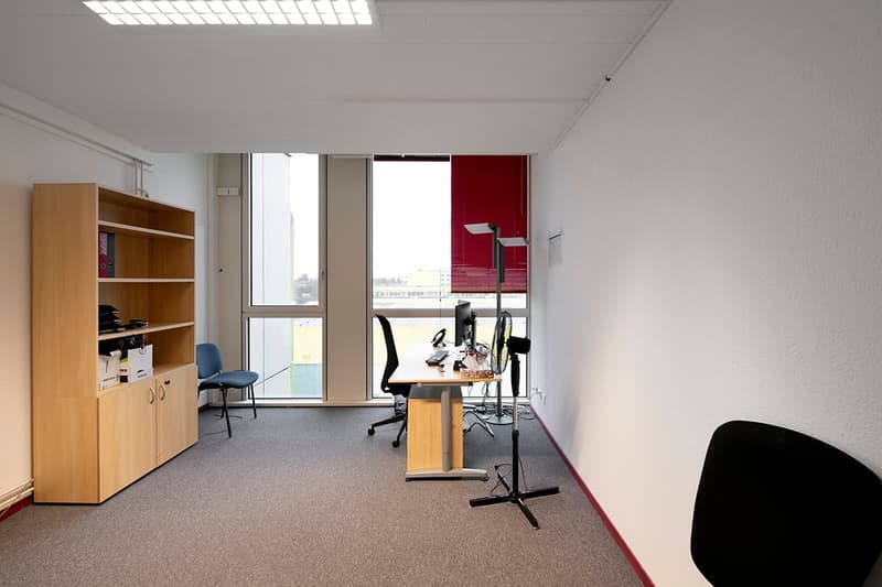 Spacious office with large windows, wooden shelving unit, desk with computer setup, office chair, and a red accent wall.