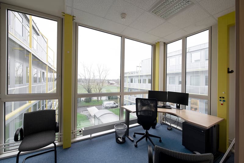 Spacious office with large windows providing a view of the outdoor scenery, including trees and buildings. The office has a desk, computer setup, and comfortable office chairs. The walls are painted in a light color scheme, and the floor is covered with b