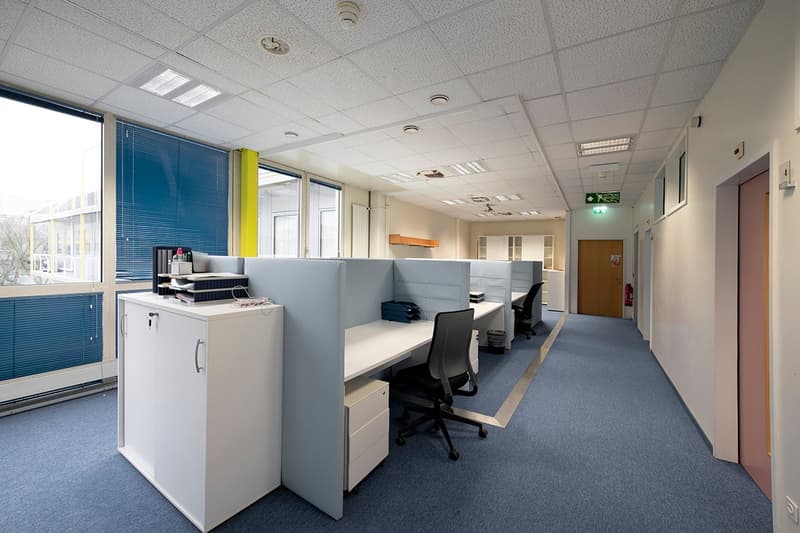 Open office space with cubicles, desks, and chairs. Bright lighting from the windows and ceiling fixtures. Refrigerator and other office equipment visible.